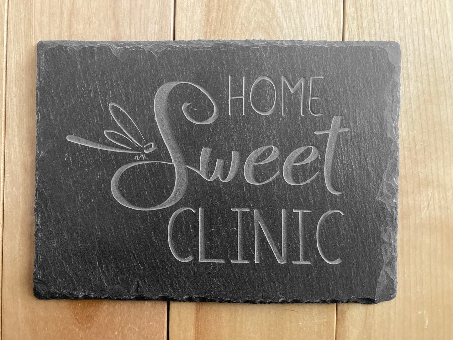 A top-down view of Home Sweet Clinic sign with dragonfly. 5"H x 7"W sand-carved black slate sign.