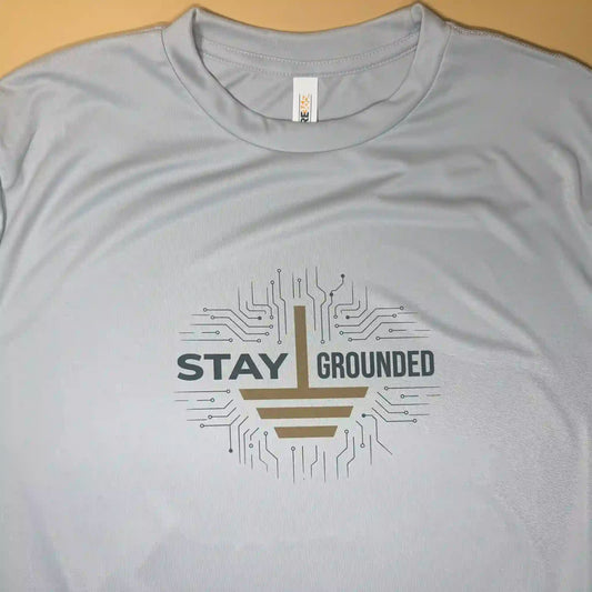 Close-up of the 'Stay Grounded' performance shirt in Platinum gray, featuring a high-contrast dark gold ground symbol and slate text sublimated into the chest and collar area.